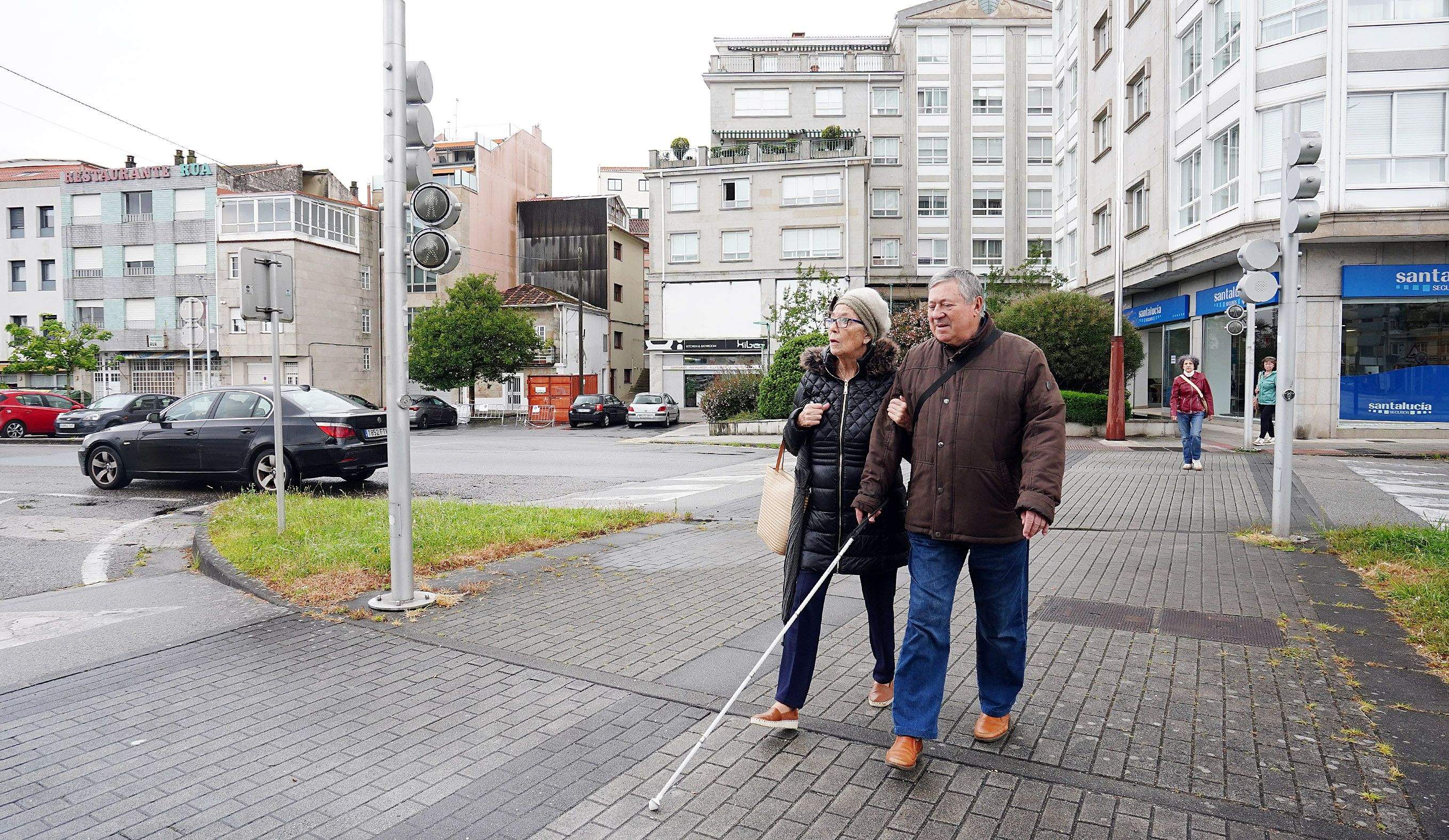 Carla e Javier cruzan o paso peonil da avenida das Corbaceiras que carece de marcas guía no firme para persoas cegas Carla e Javier cruzan o paso peonil da avenida das Corbaceiras que carece de marcas guía no firme para persoas cegas